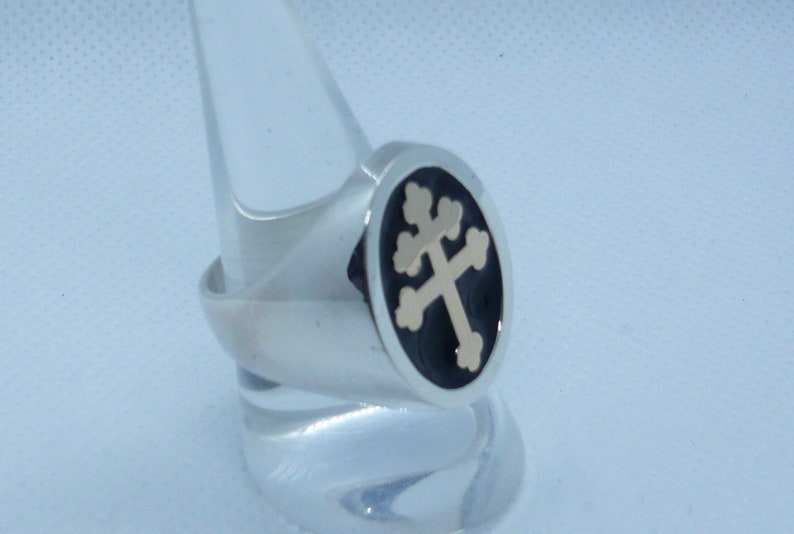 May include: A silver ring with a black enamel inlay featuring a gold-toned cross with four points. The cross is a traditional symbol of faith.