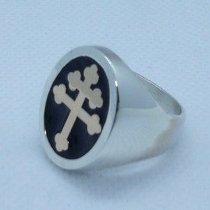 May include: A silver ring with a black oval inlay featuring a gold-colored cross with a stylized design.