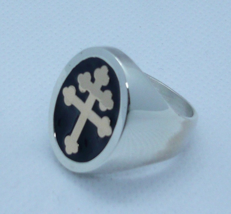 May include: A silver ring with a black oval inlay featuring a gold-colored cross with a stylized design.