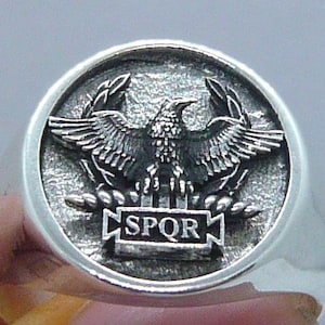 Peut inclure: Bague en argent avec un motif détaillé d'un aigle aux ailes déployées, debout sur un faisceau de blé. L'aigle est entouré d'une couronne de laurier et le texte "SPQR" est inscrit sur une bannière en dessous.