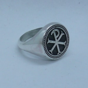 May include: A silver ring with a circular face displaying the Chi Rho symbol, a Christian emblem. The ring has a polished finish and a rope-style border. The ring is set against a light background.