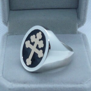 May include: A silver ring with a black and gold oval-shaped design featuring a cross. The ring is in a white velvet box.