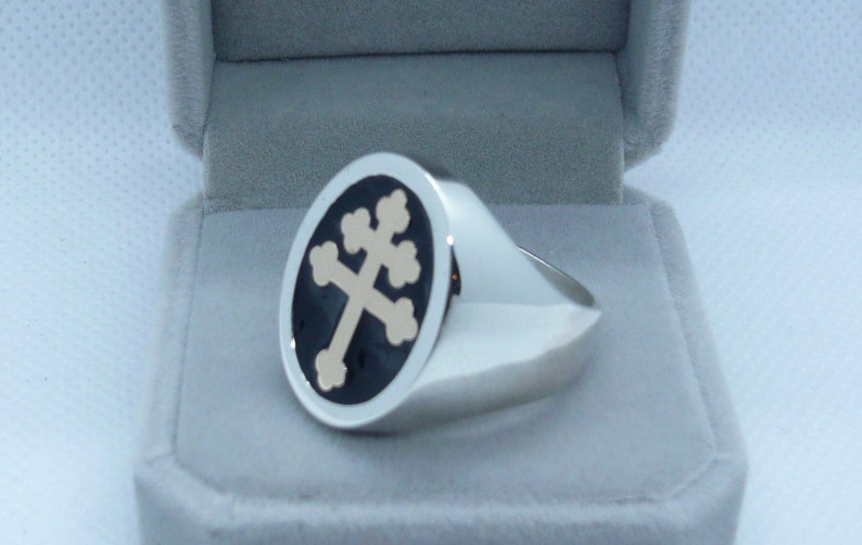May include: A silver ring with a black and gold oval-shaped design featuring a cross. The ring is in a white velvet box.