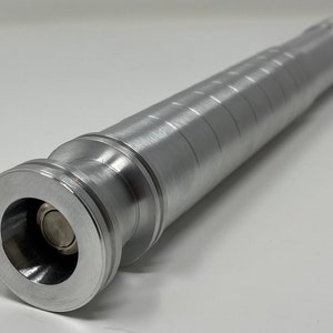 May include: A silver metal lightsaber hilt with a textured surface and a round, recessed button on the end.