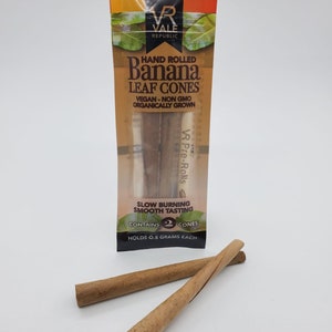 Può includere: Una confezione di coni di foglie di banana arrotolati a mano da Vale Republic. La confezione trasparente presenta dettagli arancioni e verdi, con il testo "Banana Leaf Cones". Due coni arrotolati sono mostrati davanti alla confezione. I coni sono vegani, senza OGM e coltivati ​​biologicamente.