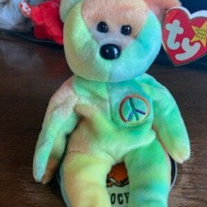 May include: A tie-dye green, blue, and pink beanie baby bear with a peace sign on its chest. The bear has black eyes and a black nose. The bear is sitting on a white surface with a red and white Ty tag.