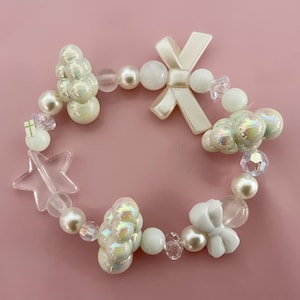 May include: A white and iridescent beaded bracelet with star, bow, and cloud charms. The bracelet is made with a variety of white beads, including pearls, iridescent beads, and clear crystal beads.