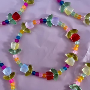May include: A colorful beaded bracelet featuring tulip-shaped beads in red, yellow, and purple, interspersed with smaller, round beads in various colors. The bracelet is set against a soft pink background.