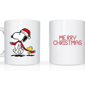 Może przedstawiać: Biały ceramiczny kubek z Snoopy w czapce Świętego Mikołaja i czerwonym szaliku, idący z Woodstockiem. Druga strona kubka ma napis "MERRY CHRISTMAS" w czerwonych literach. Świąteczny kubek.