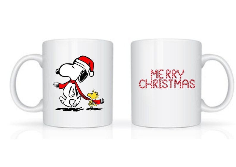 Może przedstawiać: Biały ceramiczny kubek z Snoopy w czapce Świętego Mikołaja i czerwonym szaliku, idący z Woodstockiem. Druga strona kubka ma napis "MERRY CHRISTMAS" w czerwonych literach. Świąteczny kubek.