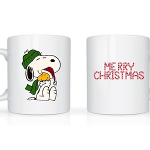 Może przedstawiać: Biały ceramiczny kubek z Snoopy w zielonej czapce i szaliku oraz czapce Świętego Mikołaja, trzymający ż&oacute;łtego ptaka. Druga strona kubka ma czerwony napis "MERRY CHRISTMAS".