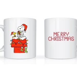 Może przedstawiać: Biały ceramiczny kubek z świątecznym wzorem. Z jednej strony Snoopy i Woodstock w czapkach Świętego Mikołaja na czerwonej budzie ozdobionej lampkami i skarpetami. Z drugiej strony napis "MERRY CHRISTMAS" w kolorze czerwonym.