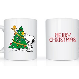 Może przedstawiać: Biały ceramiczny kubek z Snoopy dekorującym choinkę gwiazdą. Druga strona kubka ma napis "MERRY CHRISTMAS" w czerwonych, cukierkowych literach. Świąteczny prezent.