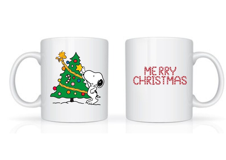 Może przedstawiać: Biały ceramiczny kubek z Snoopy dekorującym choinkę gwiazdą. Druga strona kubka ma napis "MERRY CHRISTMAS" w czerwonych, cukierkowych literach. Świąteczny prezent.