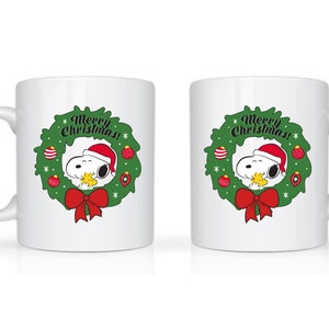 Może przedstawiać: Biały ceramiczny kubek z Snoopy w czapce Świętego Mikołaja, otoczony zielonym wieńcem z czerwonymi ozdobami i czerwoną kokardą. Tekst "Merry Christmas!" jest wyświetlany nad Snoopy. Świąteczny, świąteczny design.