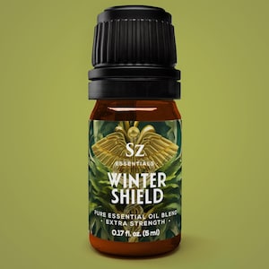 Puede incluir: Una pequeña botella de vidrio ámbar con tapa negra, etiquetada como "SZ Essentials Winter Shield". La etiqueta presenta un emblema dorado y follaje verde, con texto que indica una mezcla de aceite esencial puro, extra fuerte y 5 ml.