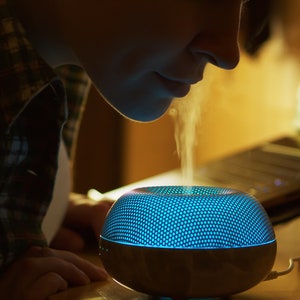 Puede incluir: Una persona inhala el vapor de un difusor de aceites esenciales azul y gris con un patr&oacute;n de panal en la parte superior.