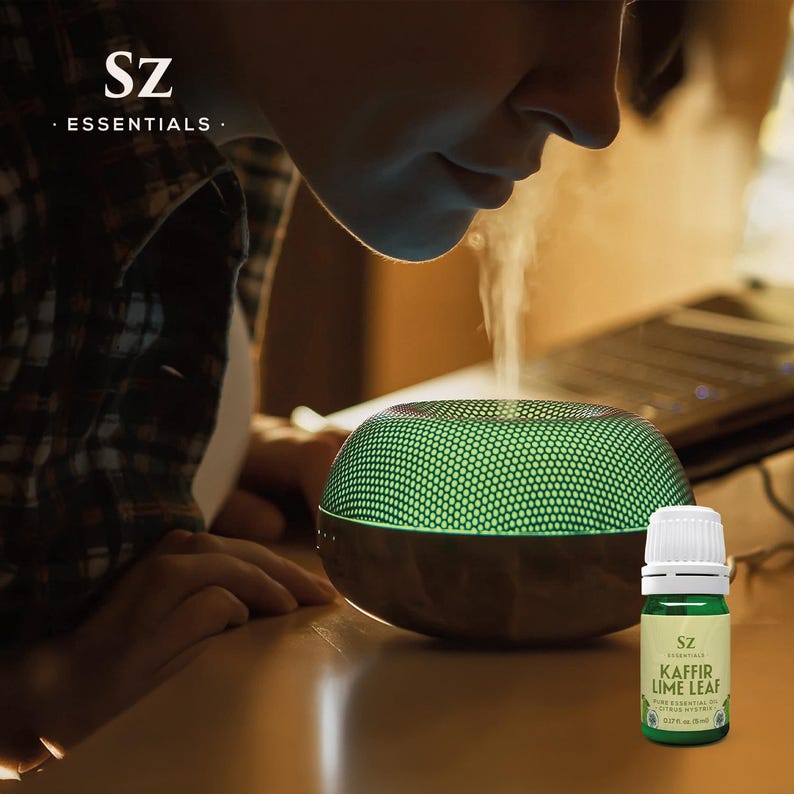 May include: A person inhales the aroma of Kaffir Lime Leaf essential oil diffusing from a green and brown diffuser. The bottle of essential oil is labeled "SZ Essentials Kaffir Lime Leaf Pure Essential Oil 10 ml / 0.33 fl oz"