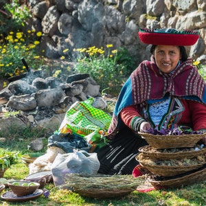 Puede incluir: Una mujer con un sombrero y ropa tradicionales de color rojo y azul est&aacute; sentada en el suelo con una pila de cestas tejidas llenas de hierbas secas.