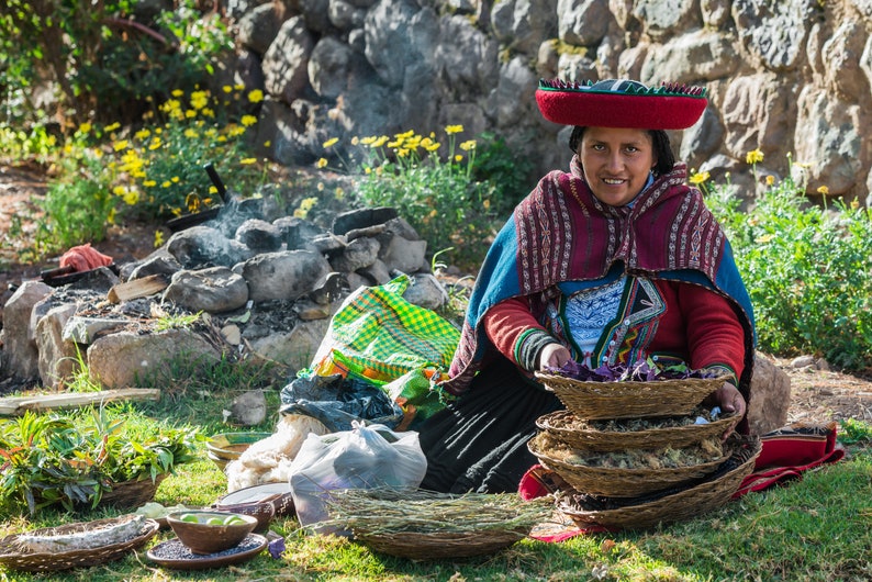 Puede incluir: Una mujer con un sombrero y ropa tradicionales de color rojo y azul est&aacute; sentada en el suelo con una pila de cestas tejidas llenas de hierbas secas.