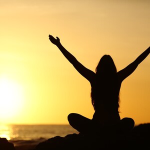May include: Silhouette of a person with arms raised in the air, sitting with legs crossed, against a bright orange sunset over the ocean.