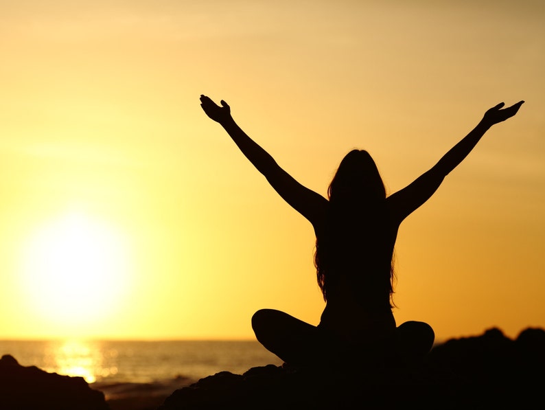 May include: Silhouette of a person with arms raised in the air, sitting with legs crossed, against a bright orange sunset over the ocean.