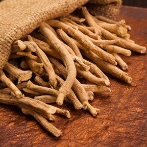 May include: A burlap sack filled with dried, light brown, thin, and long herbal roots. The roots are spilling out of the sack onto a dark brown wooden surface.