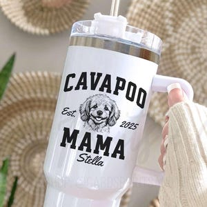 May include: White insulated tumbler with a handle, featuring the text "CAVAPOO MAMA Stella" and a Cavapoo dog illustration. The tumbler includes a straw and a silver lid. The design includes the year "2025" and the word "Est."