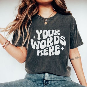 May include: A dark grey t-shirt with a white graphic that says "YOUR WORDS HERE" in a retro font. The shirt has a relaxed fit and is made of a soft, comfortable fabric.