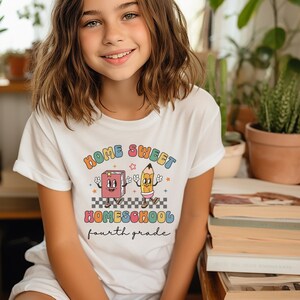 May include: A white t-shirt with a colorful graphic design that says "Home Sweet Homeschool fourth grade". The graphic features a cartoon book and pencil holding hands.