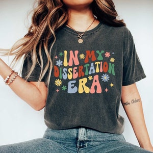 May include: A dark gray t-shirt with the text "IN MY DISSERTATION ERA" in colorful, retro-style lettering. The text is surrounded by small flowers and stars.