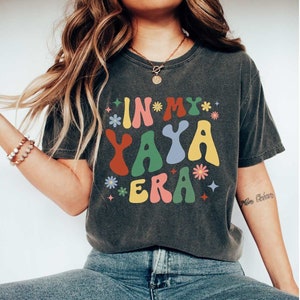 Puede incluir: Una camiseta gris oscuro con un gr&aacute;fico colorido que dice "In My Yaya Era" con flores y estrellas. La camiseta la lleva puesta una persona con el pelo largo y casta&ntilde;o y unos vaqueros azules.