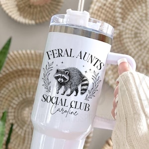 May include: White stainless steel travel mug with a black and white raccoon graphic and the text "Feral Aunts Social Club Caraline".