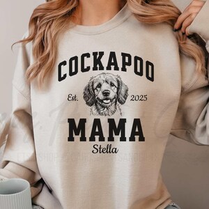 May include: A beige sweatshirt with a black graphic of a Cockapoo dog. The graphic says "Cockapoo" in a black arched font, "Est. 2025" in smaller black font, "Mama" in large black font, and "Stella" in smaller black font.