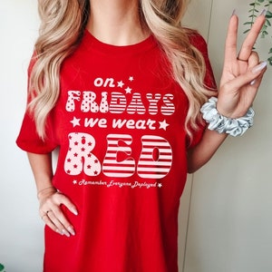 Peut inclure: T-shirt rouge avec un motif blanc qui dit "on Fridays we wear red" avec des &eacute;toiles et des rayures. Le texte "Remember Everyone Deployed" est sous le motif.