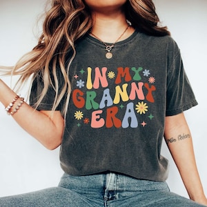 May include: A dark gray t-shirt with the text "IN MY GRANNY ERA" in colorful retro font with flowers and stars around the text.