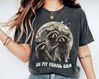 May include: A dark gray t-shirt with a graphic of three raccoons in front of a full moon with stars. The text "In My Feral Era" is printed below the raccoons.