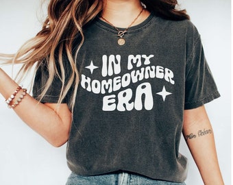Könnte beinhalten: Eine Frau trägt ein dunkelgraues T-Shirt mit dem Text "In My Homeowner Era" in Weiß. Sie trägt eine blaue Jeans und weiße Turnschuhe.