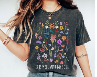 May include: A charcoal gray short-sleeve t-shirt featuring a colorful botanical print of various wildflowers and small insects. The text "IT IS WELL WITH MY SOUL" is printed in white capital letters below the floral graphic design.