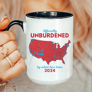 May include: White ceramic mug with a black rim and handle. The mug features a red, white, and blue map of the United States with the text "Officially Unburdened by what has been 2024".