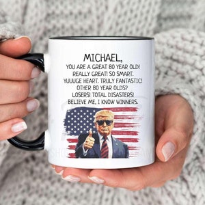 May include: White ceramic coffee mug with a black rim and handle. The mug features a graphic of a man with a thumbs up, an American flag, and the text "MICHAEL, YOU ARE A GREAT 80 YEAR OLD! REALLY GREAT! SO SMART. YUUUGE HEART. TRULY FANTASTIC! OTHER 80 YEAR OLDS? LOSERS! TOTAL DISASTERS! BELIEVE ME, I KNOW WINNERS."