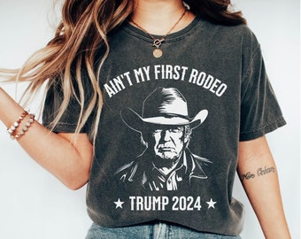 May include: A dark gray t-shirt with a white graphic of a man wearing a cowboy hat and the text "Ain't My First Rodeo" and "Trump 2024".
