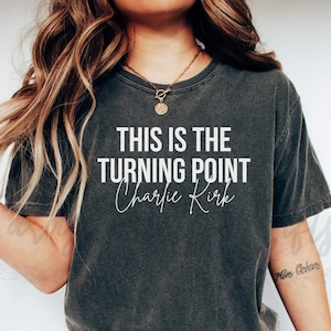 May include: Dark gray t-shirt with the text "THIS IS THE TURNING POINT Charlie Kirk" in white. The shirt is a crew neck style and appears to be made of a soft, comfortable material. The shirt is worn by a person.
