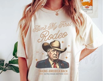 May include: Ivory-colored t-shirt with the text "Ain't My First Rodeo" and an illustration of a man wearing a cowboy hat. The graphic includes the text "Taking America Back" and "Ultra MAGA Edition".
