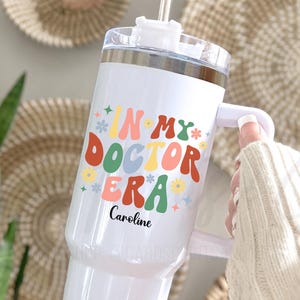 May include: White tumbler with a handle and a straw, featuring the text "IN MY DOCTOR ERA" in colorful, retro-style lettering, with the name "Caroline" below. The tumbler has a silver lid.
