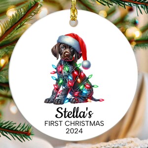 May include: A white ceramic Christmas ornament featuring a brown and black puppy wearing a red Santa hat and tangled in colorful Christmas lights. The ornament reads "Stella's First Christmas 2024".