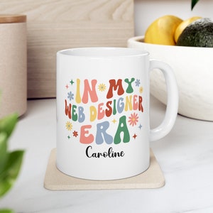 May include: A white ceramic mug featuring the colorful text "IN MY WEB DESIGNER ERA" in a wavy, retro-inspired font, accented by small flower and star icons. The name "Caroline" is printed in a black script font below the main text.