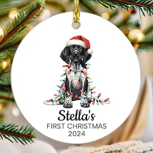 May include: A white ceramic Christmas ornament featuring a black and white dog wearing a Santa hat and wrapped in colorful Christmas lights. The ornament reads "Stella's First Christmas 2024". A gold string is attached for hanging.
