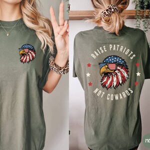 May include: A green t-shirt with a graphic of an eagle wearing sunglasses and a red, white, and blue bandana. The text "Raise Patriots Not Cowards" is printed on the back of the shirt.
