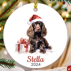 May include: A Christmas ornament featuring a brown and white dog wearing a red Santa hat and sitting next to a wrapped gift. The ornament is white with the name "Stella" and the year "2024" printed on it.